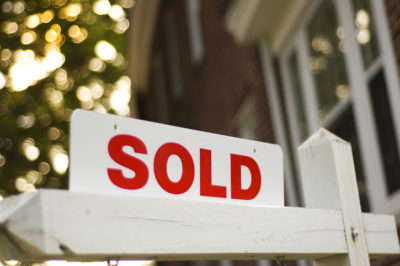 Real Estate "sold" sign with red brick building and trees blurry in the background